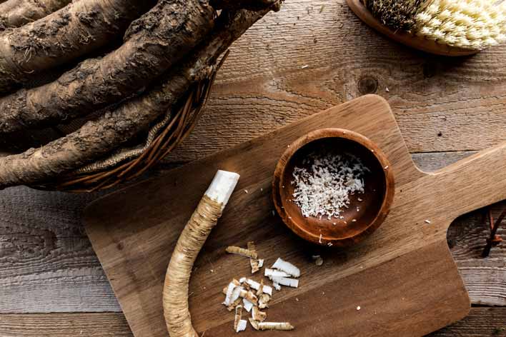 Frisch geriebener Meerrettich auf einem Holzbrett. Im Hintergrund Meerrettichwurzeln und eine Bürste
