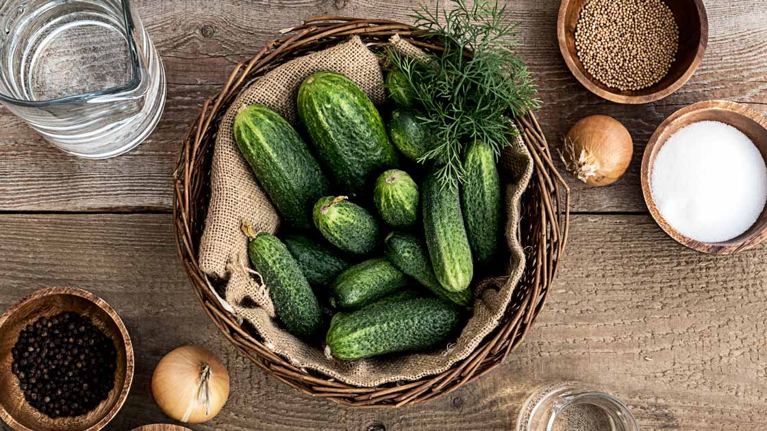  Die benötigten Zutaten für Blitzgurken liegen auf einem Holztisch: Salz, Gewürze, Einlegegurken, Zwiebeln, Pfeffer