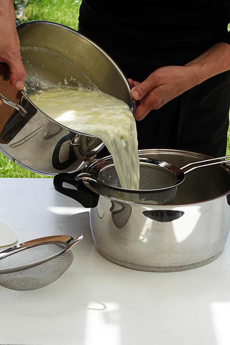 Eine Frau schüttet einen Topf mit Käsebruch durch ein Sieb in einen anderen Topf um den Käsebruch von der Molke zu trennen