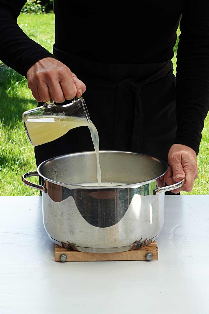 Milch wird in einem Topf erhitzt. Eine Frauenhand gießt Zitronensatz in die Milch wodurch diese gerinnt.
