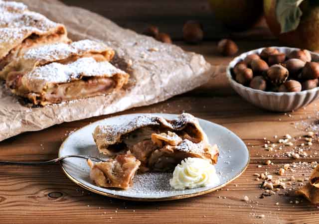 Ein frisch gebackener Apfelstrudel in Scheiben geschnitten. Ein Stück Apfelstrudel liegt auf einem Teller. 