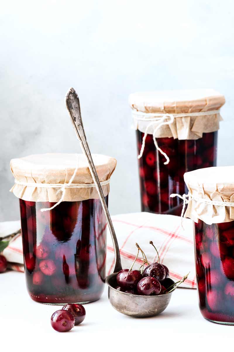 Drei Einmachgläsern gefüllt mit Kirschen. Dazwischen ein Schöpflöffel gefüllt mit frischen Kirschen