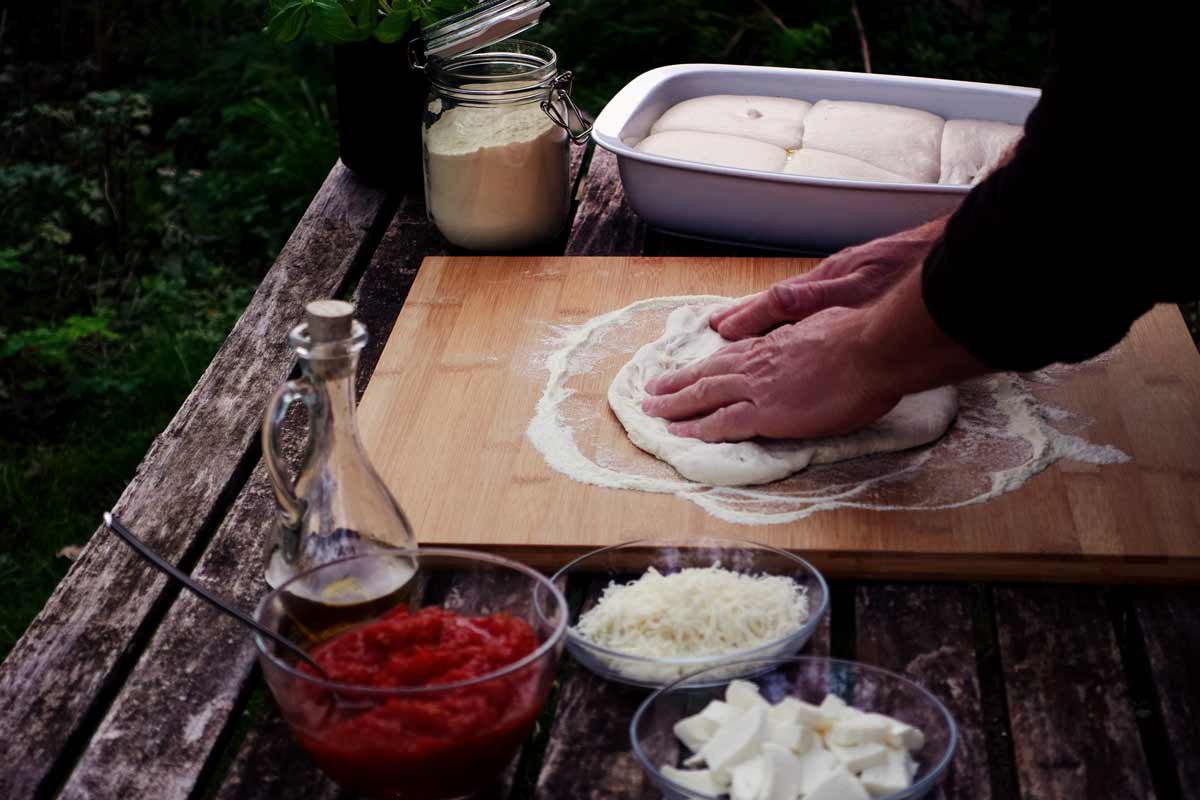 Ein Mann formt die neapolitanische Pizza traditionell mit den Händen. An der Seite stehen die Zutaten griffbereit.