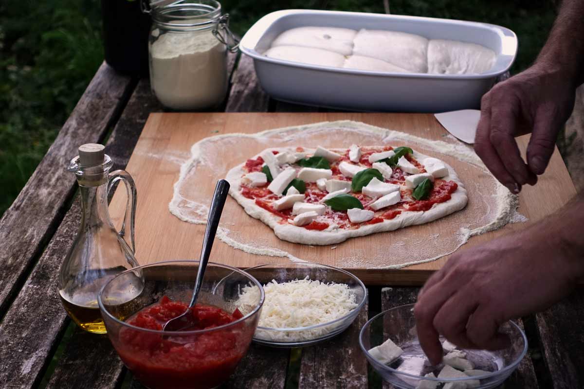 Ein Mann belegt eine  neapolitanische Pizza mit den traditionellen Zutaten: Tomatensauce, Mozarella Und Basilikum. An der Seite stehen alle Zutaten griffbereit.