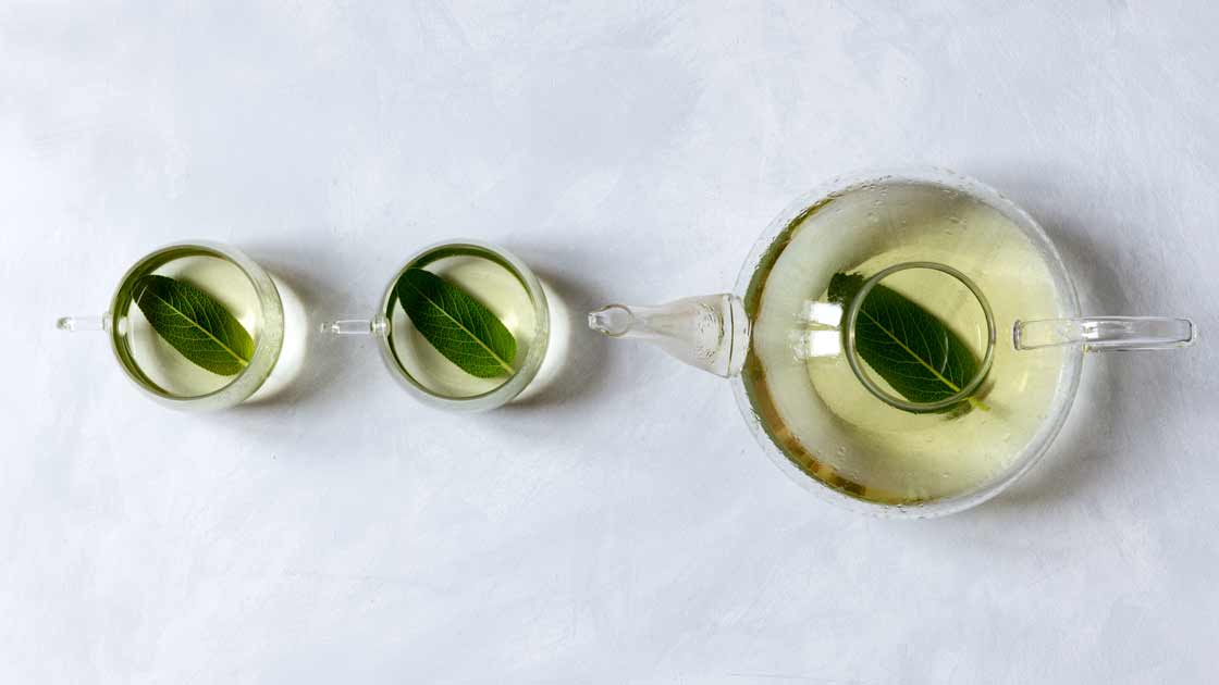 Gläserne Teekanne und zwei Teetassen mit frisch gebrühtem Salbeitee und jeweils einem Salbeiblatt