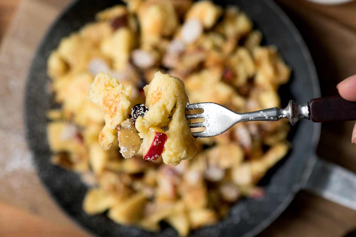 Lockerer, fluffiger Kaiserschmarrn auf einer Gabel. Im Hintergrund eine Portion Kaiserschmarrn mit Äpfeln, Nüssen und Rosinen in einer Eisenpfanne