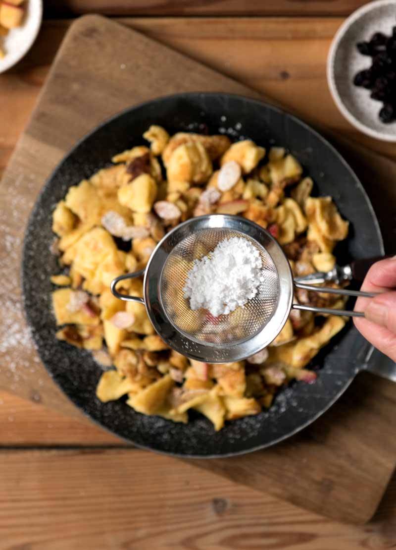 Eine Hand stäubt Puderzucker über eine Pfanne mit Kaiserschmarrn
