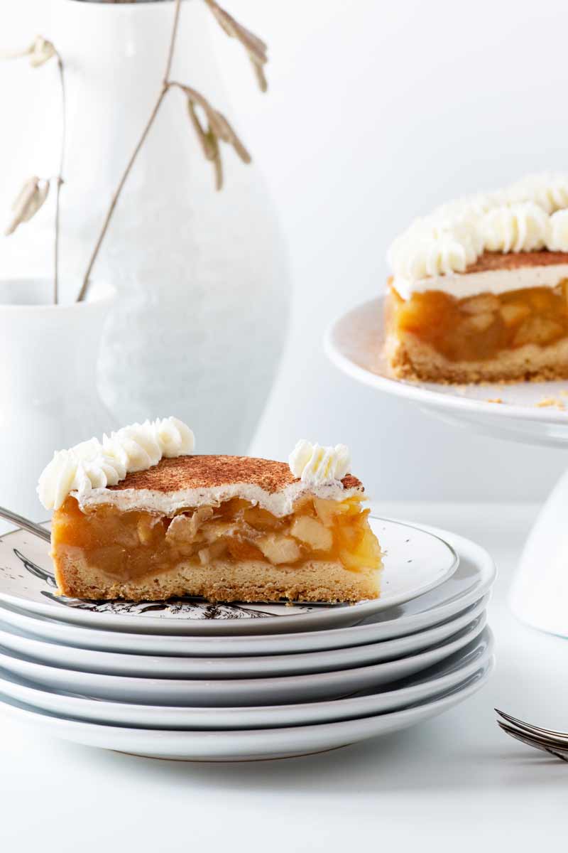 Ein Stück weihnachtliche, schwedische Apfeltorte dekoriert mit Sahne und Zimtzucker auf einem Stapel Teller.
