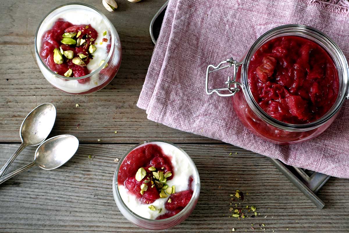 Rhabarberkompott mit Joghurt und gehackten Pistazien in Glasschalen, daneben ein Weckglas mit eingekochtem Kompott