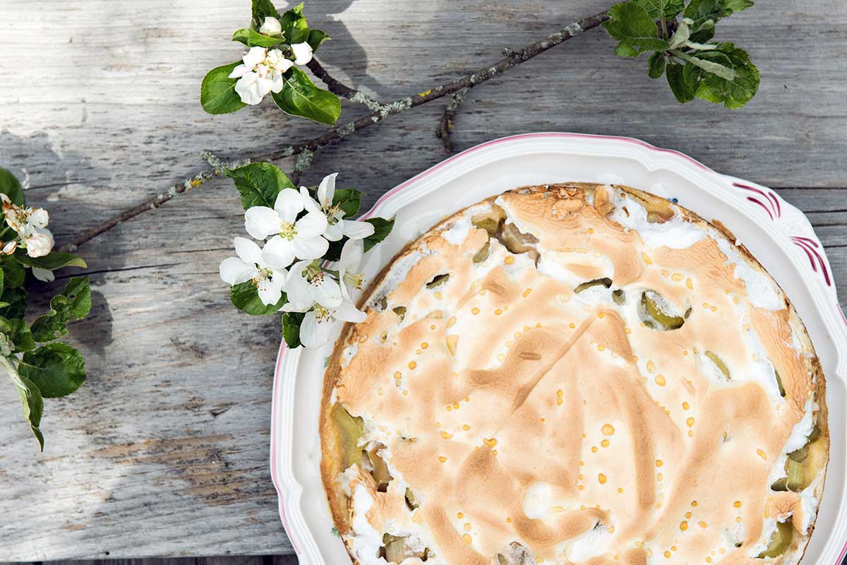 Rhabarberkuchen mit goldbrauner Baiserhaube in einer runden Backform, dekoriert mit Apfelblütenzweigen auf rustikalem Holztisch